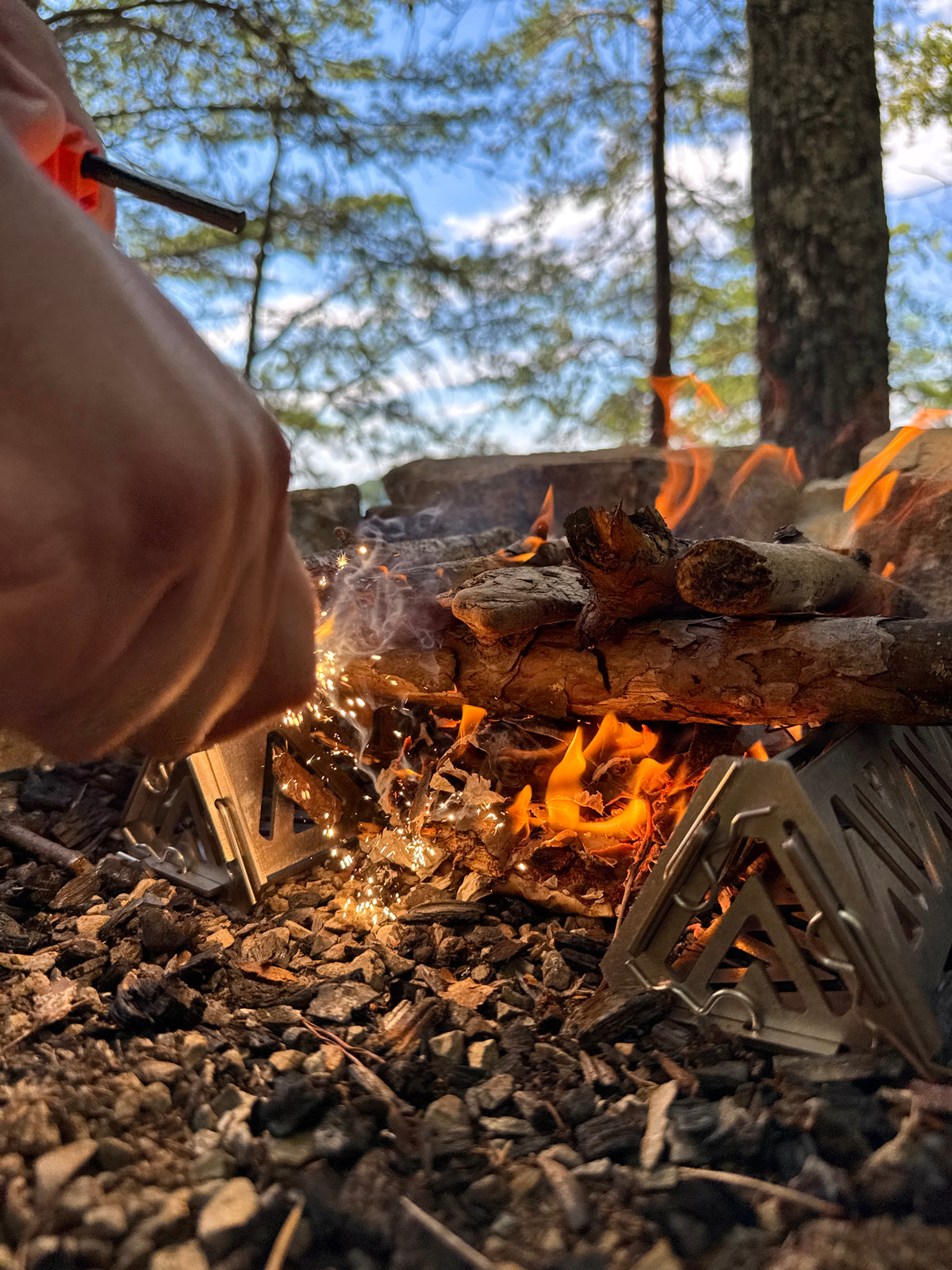Person using a fire starter to ignite wood in a campfire with trees in the background