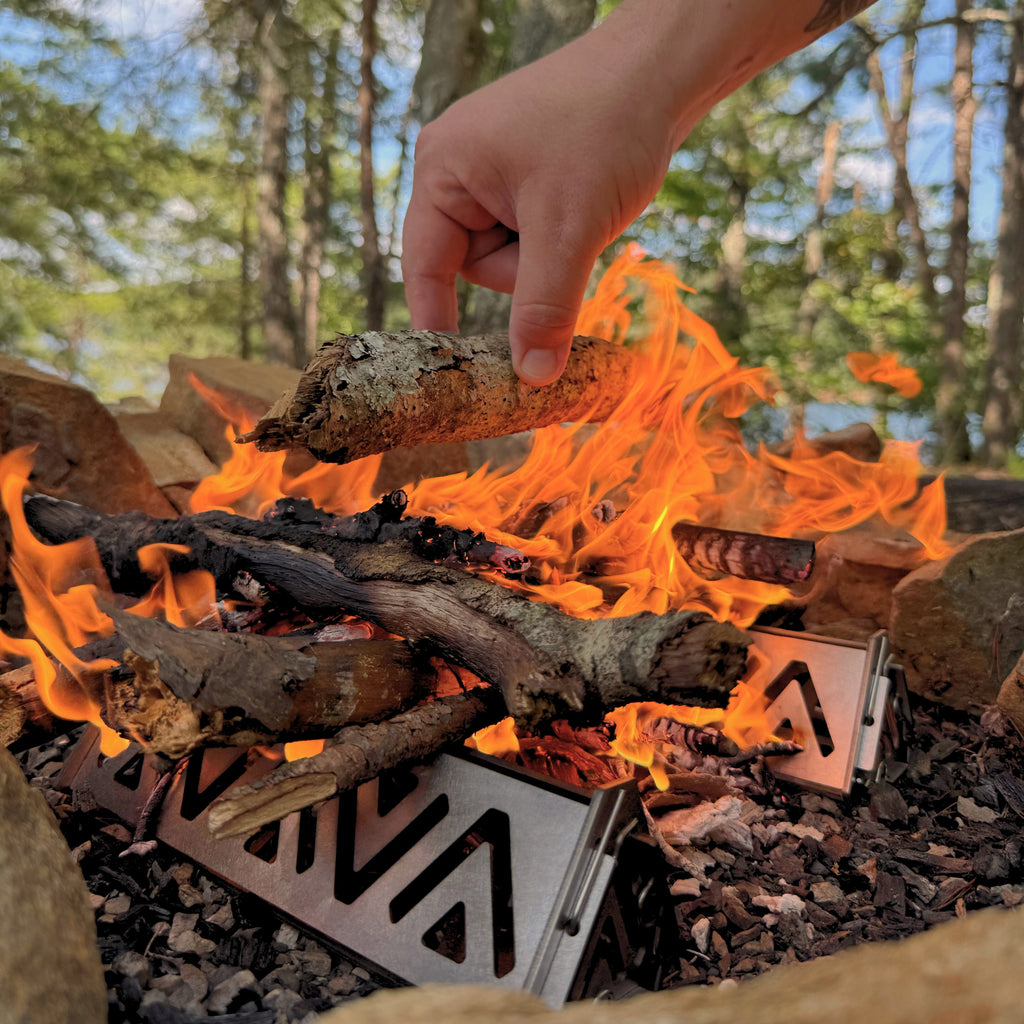 Person adding wood to a campfire with a metal fire pit in the foreground, surrounded by trees.