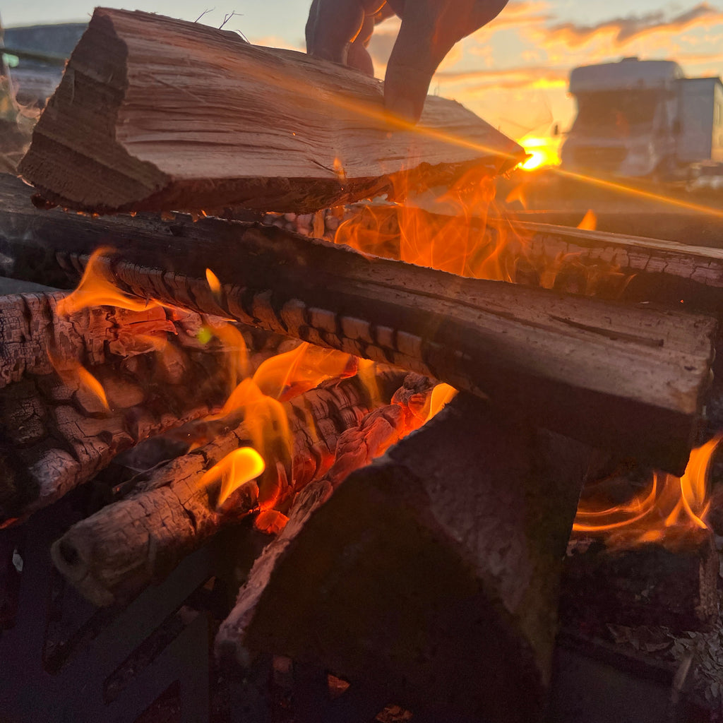 Person holding kindling over a fire with flames and smoke in the background