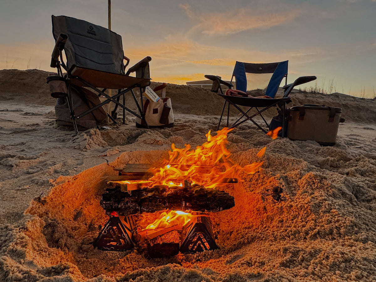 Campfire on a sandy beach with two folding chairs and a cooler in the background.